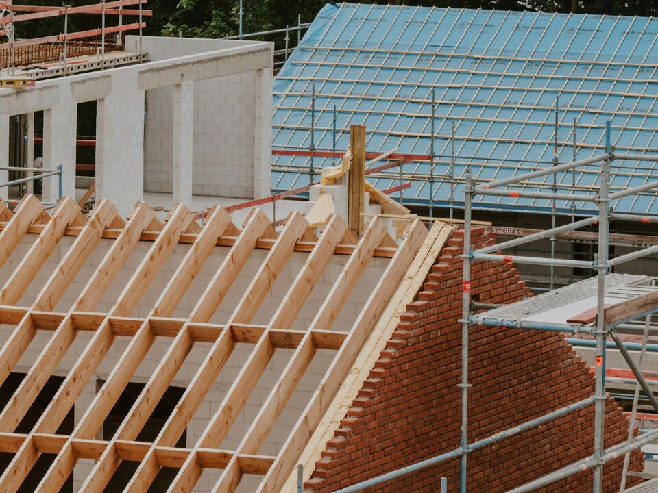 Maison en construction : chevrons et lattes en bois, pignon en briques, sous-toiture bleue et échafaudage.
