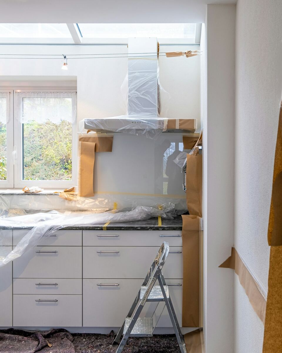 Cuisine en cours de rénovation : meubles et plan de travail protégés par des bâches et du papier, escabeau au sol.