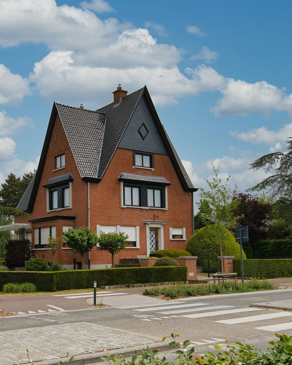 Maison en briques rouges à haute toiture d’ardoises, entourée d’un jardin, sous un ciel bleu.