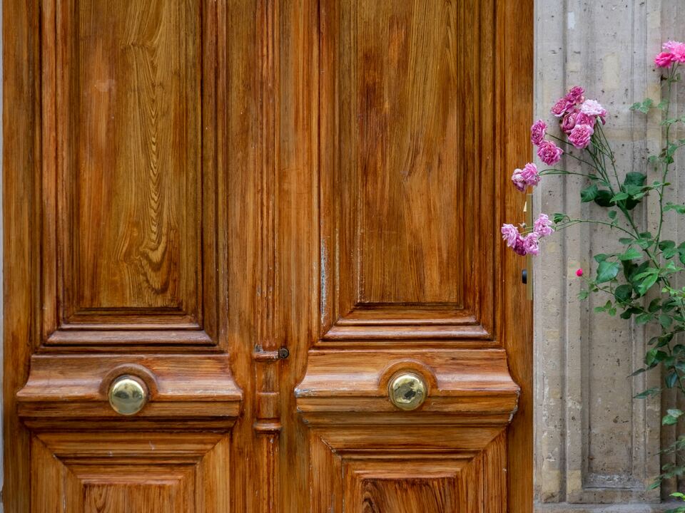 Porte d’entrée double en bois verni, poignées rondes en laiton, rosier grimpant contre la façade.