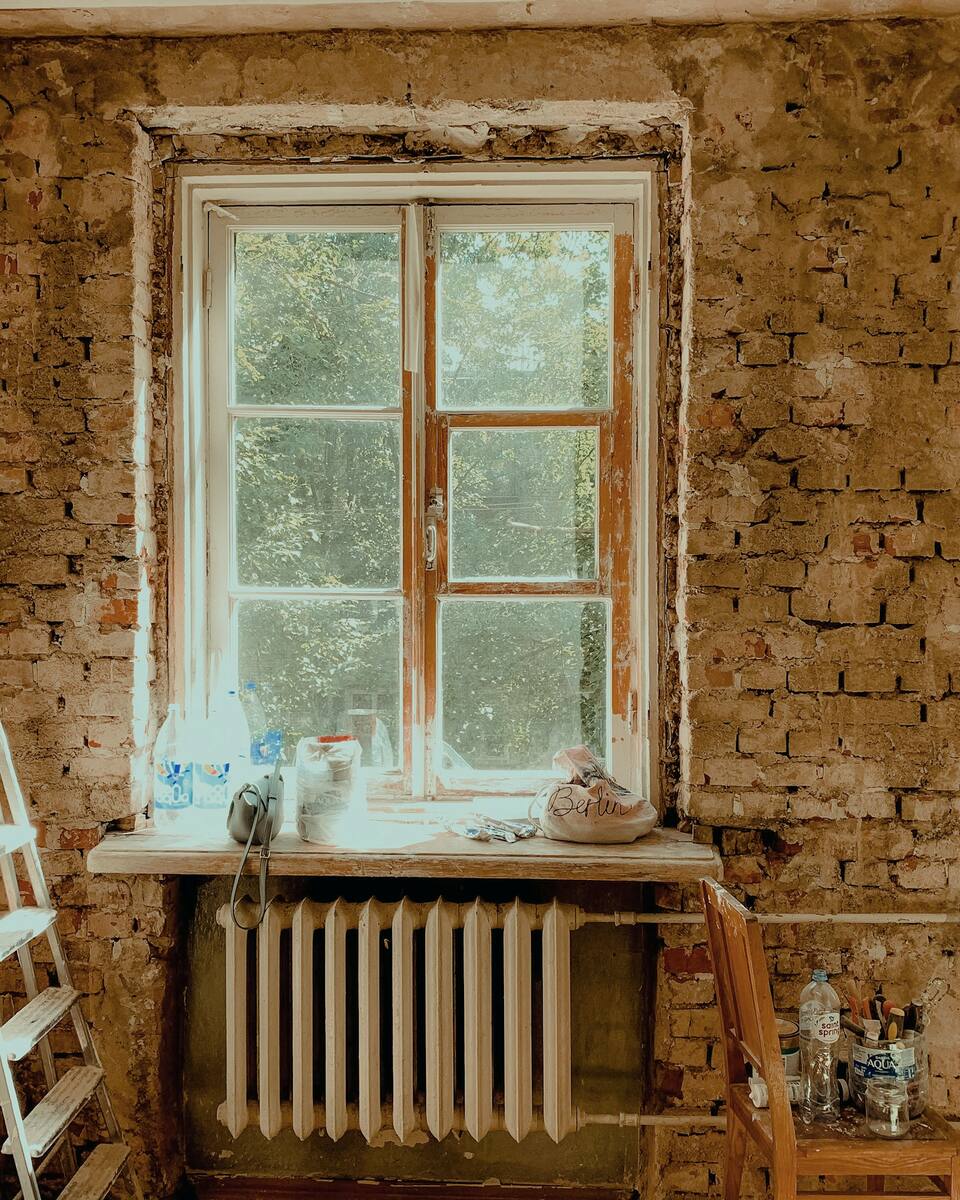 Mur de briques décapé autour d’une fenêtre ancienne à croisillons, escabeau et radiateur : une pièce en pleine rénovation.