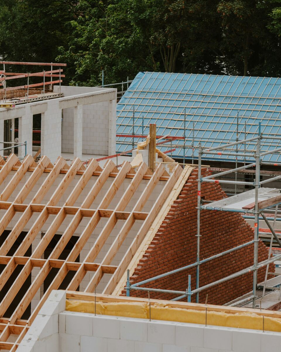 Maison en construction : chevrons et lattes en bois, sous-toiture bleue, échafaudage et murs en blocs.
